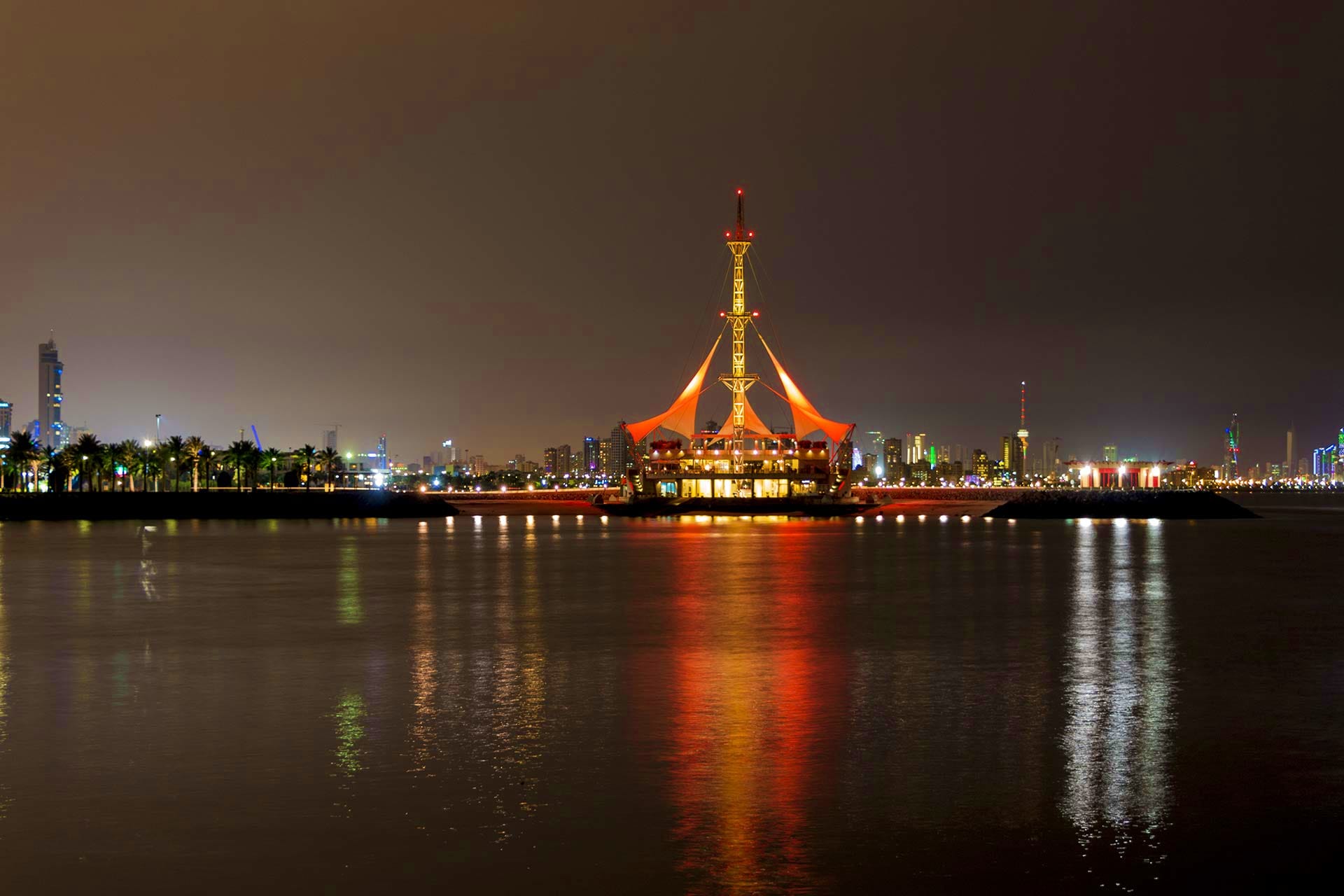 Illuminated golden architectural landmark with orange tent-like structure reflecting in dark water at night, palm trees and c